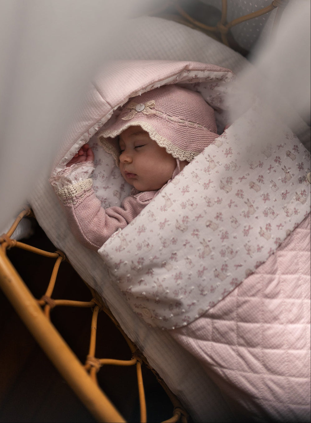 A sleeping newborn baby wrapped in a light pink floral blanket and wearing a matching pink bonnet, lying in a wooden bassinet under soft white drapery.