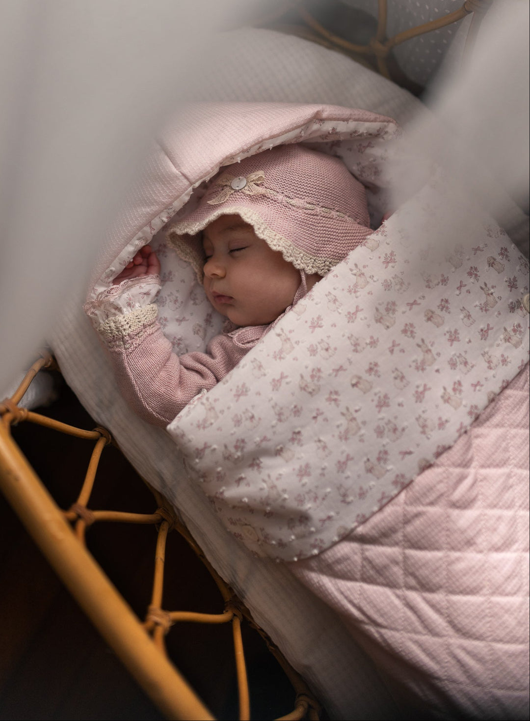 A sleeping newborn baby wrapped in a light pink floral blanket and wearing a matching pink bonnet, lying in a wooden bassinet under soft white drapery.