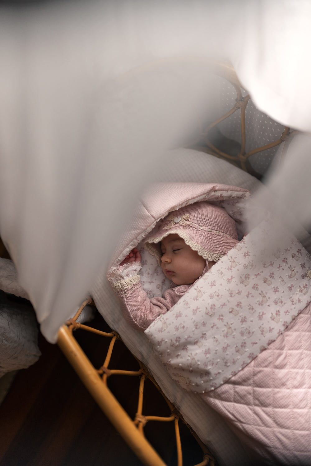 A sleeping newborn baby wrapped in a light pink floral blanket and wearing a matching pink bonnet, lying in a wooden bassinet under soft white drapery.