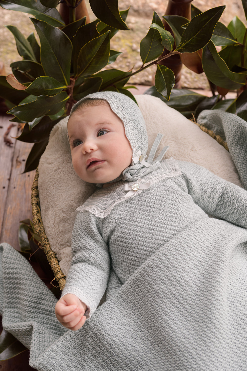 A baby wearing a textured light green knit bonnet and matching long-sleeve romper, lying on a beige cushion outdoors with green leaves in the background.