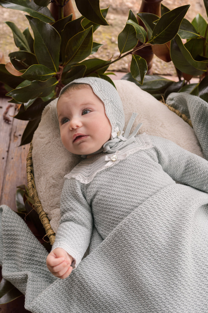 A baby wearing a textured light green knit bonnet and matching long-sleeve romper, lying on a beige cushion outdoors with green leaves in the background.