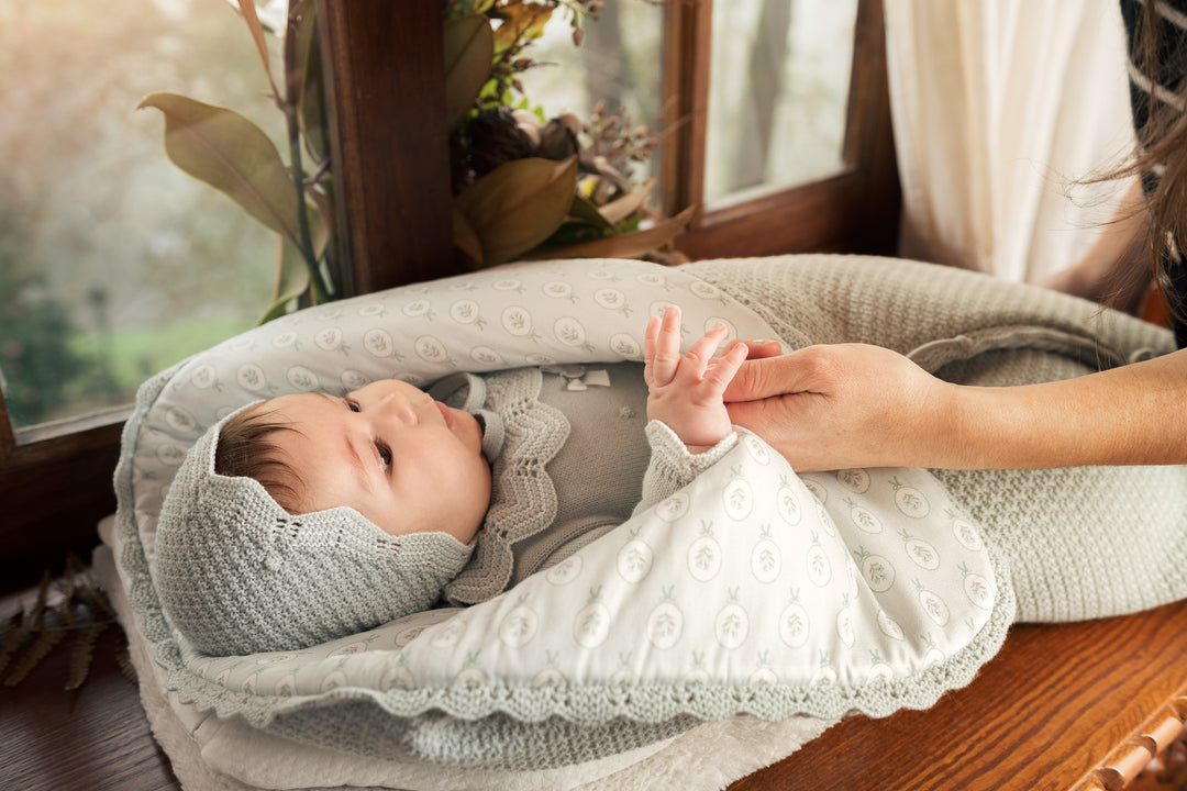 A baby wrapped in a soft white patterned swaddle blanket and wearing a matching light gray knit bonnet, resting in a gray baby lounger on a wooden surface near a window, with an adult hand holding the baby's hand.