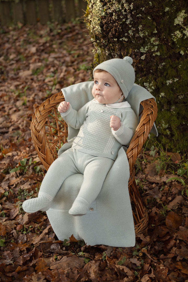 A baby wearing a light sage green knit outfit with a matching beanie sits in a woven wicker chair outdoors on a bed of brown autumn leaves, with a mossy tree trunk in the background.
