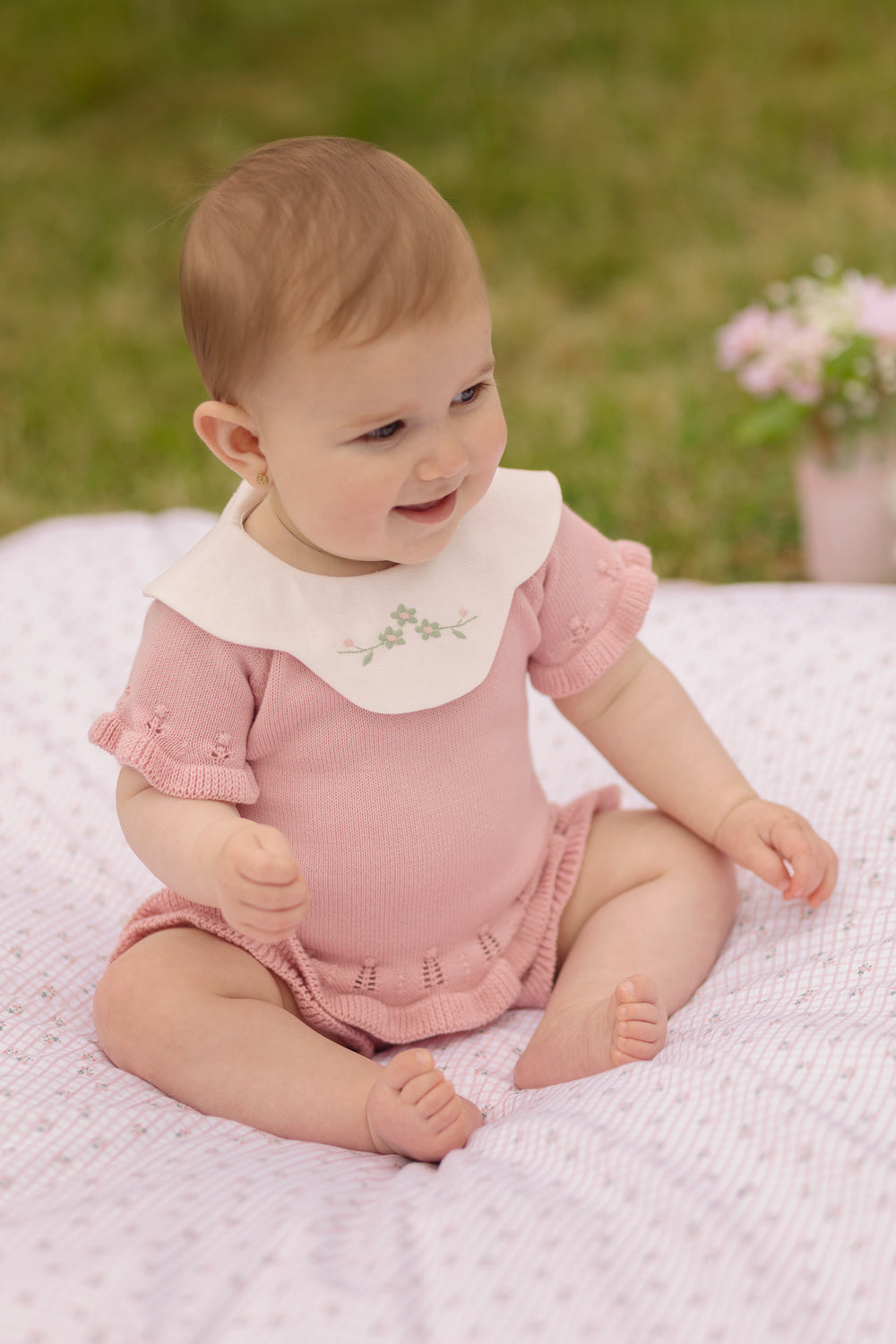 Baby in a pink outfit with a white collar sitting on a blanket outdoors.