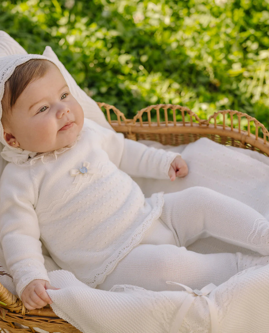 Baby in a white outfit sitting in a wicker basket with greenery in the background