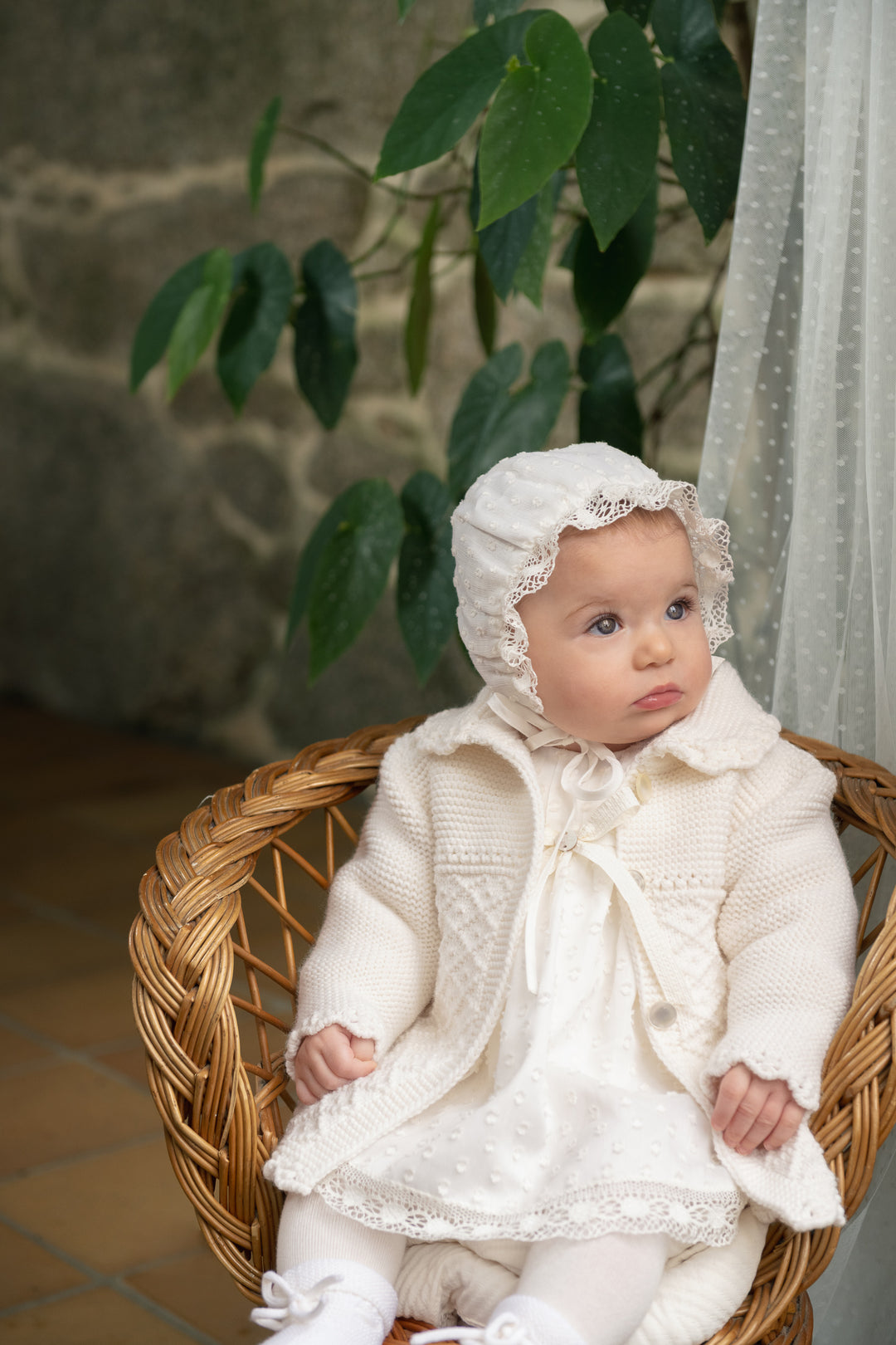 A baby wearing a cream-colored knit bonnet and matching knitted outfit sits in a woven wicker chair, with green leaves and a stone wall in the background.