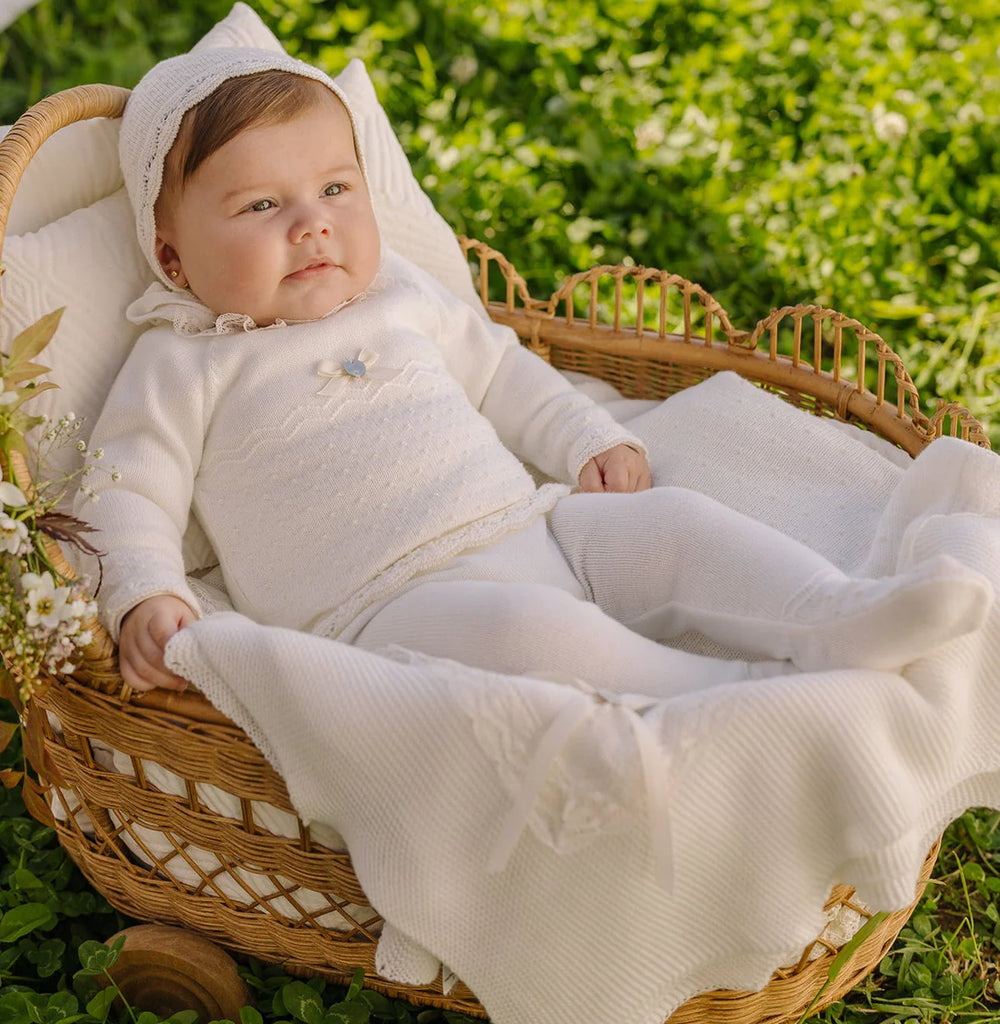 Baby in a white outfit sitting in a wicker basket outdoors.