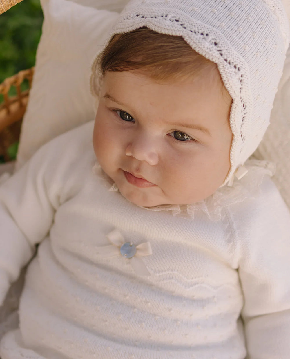 Baby wearing a white outfit with a lace bonnet, sitting on a soft surface.
