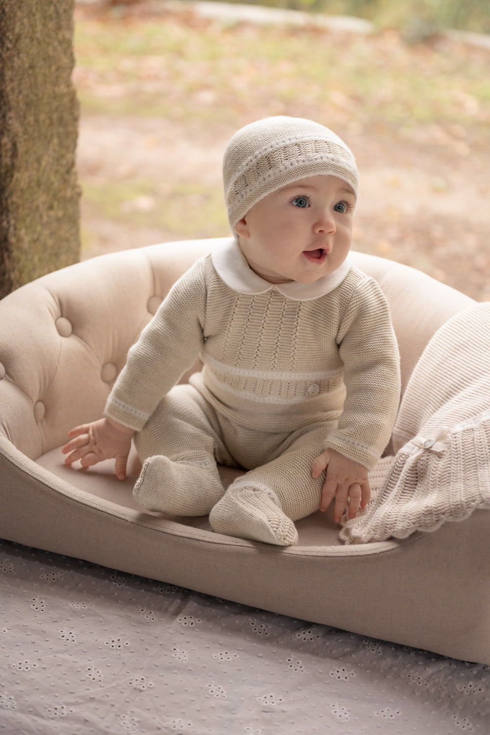 A baby wearing a cream-colored knit one-piece outfit with a matching beanie sits in a tufted cream baby chair outdoors.