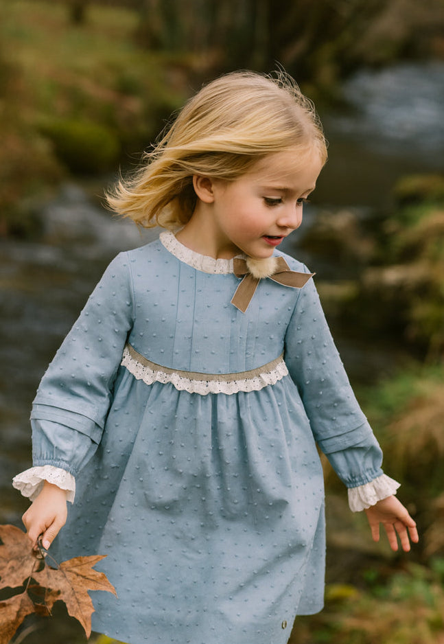 A young girl with blonde hair wearing a dusty blue long-sleeve dress with white lace trim and a brown bow, standing outdoors near a stream.