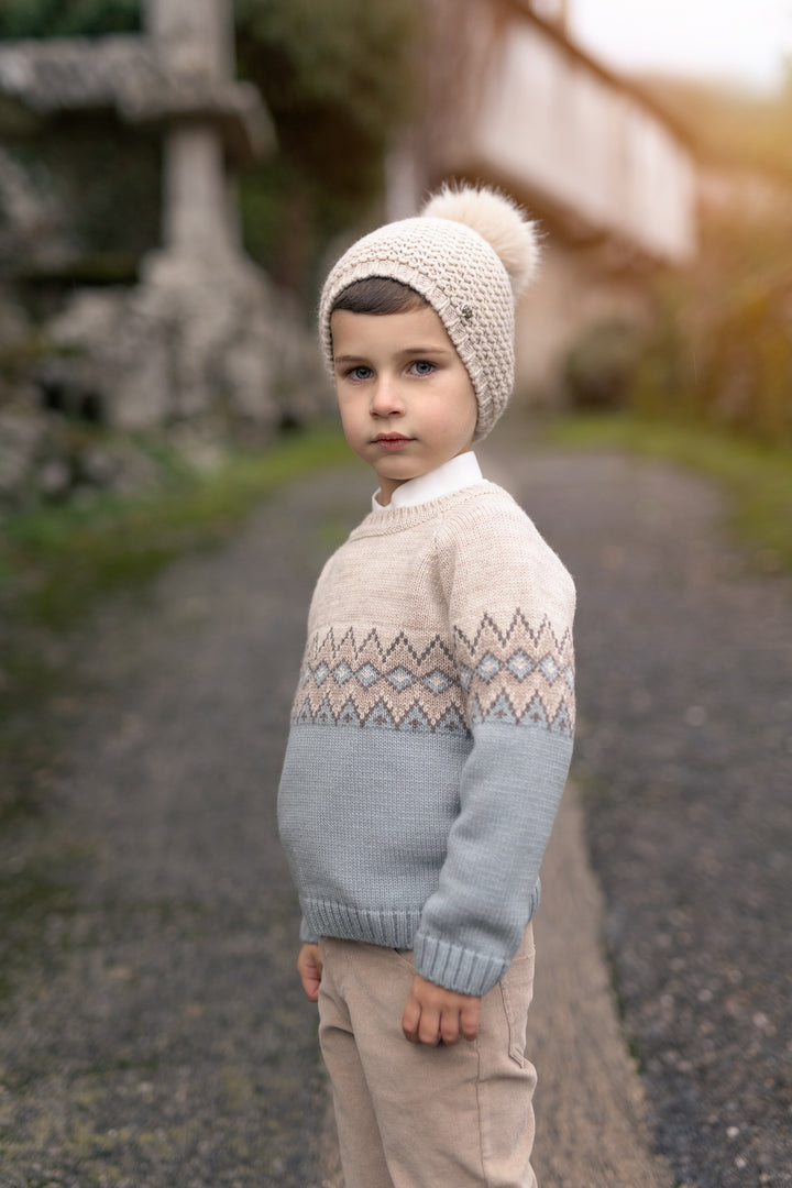 A young boy wearing a beige knit sweater with a geometric pattern across the chest, a light blue lower section, and a matching beige knit beanie with a pom-pom, standing on an outdoor gravel path.