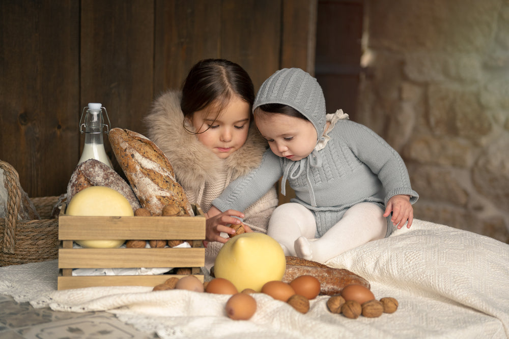 Two young children sitting on a cream blanket surrounded by rustic bread, eggs, walnuts, and a wooden crate in a warm, cozy indoor setting.