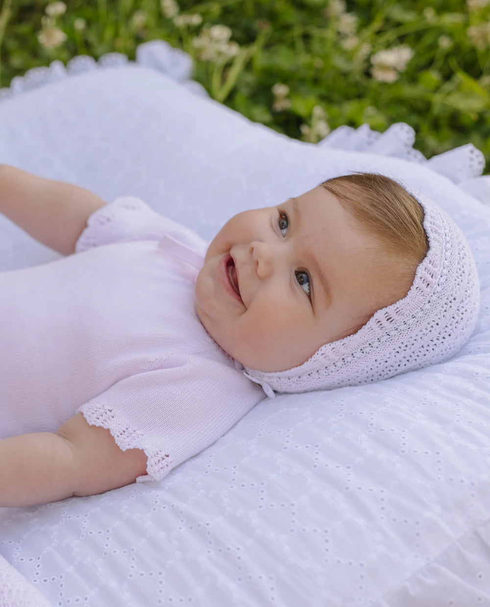 Baby lying on a white blanket outdoors with greenery in the background