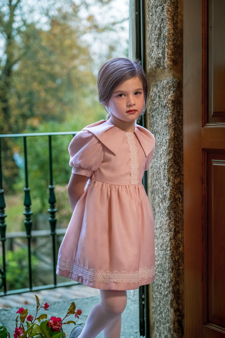 A young girl with brown hair stands by a stone pillar and glass door, wearing a light pink short-sleeved dress with lace trim and white tights.