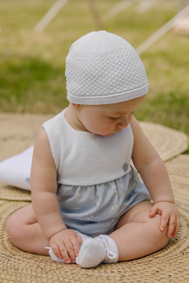 Baby sitting on a woven mat wearing a white textured hat and light blue outfit.