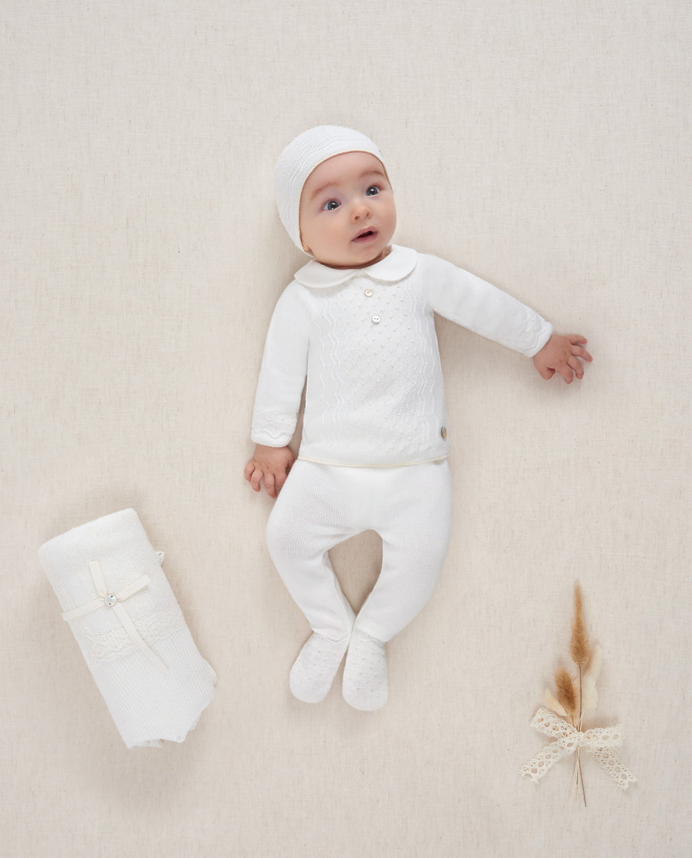 Baby in white outfit lying on a beige surface with a white bootie beside.
