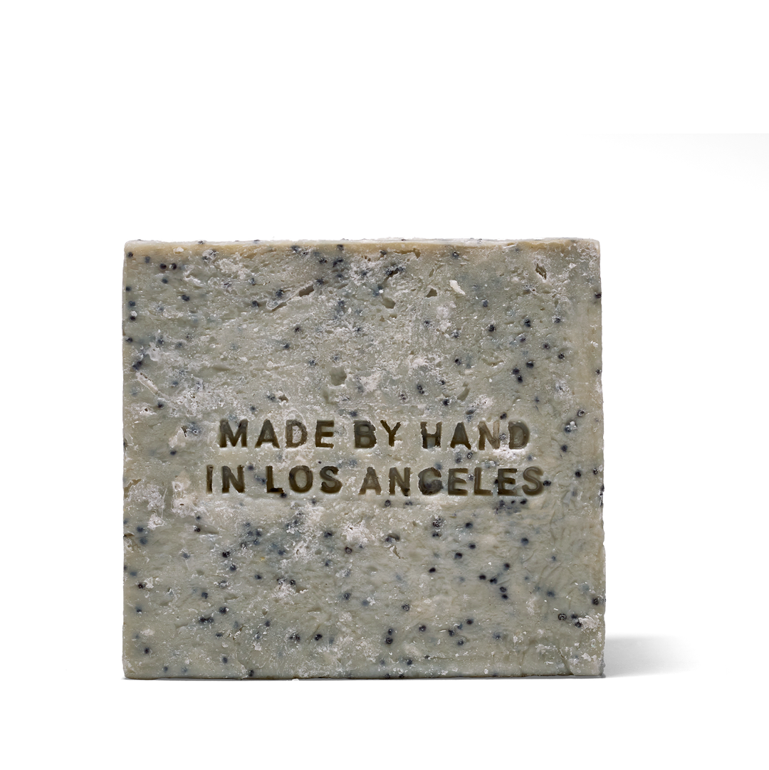 A rectangular bar of speckled soap in a light gray-green tone with dark flecks, featuring the embossed text 'MADE BY HAND IN LOS ANGELES' on the front, set against a plain white background.