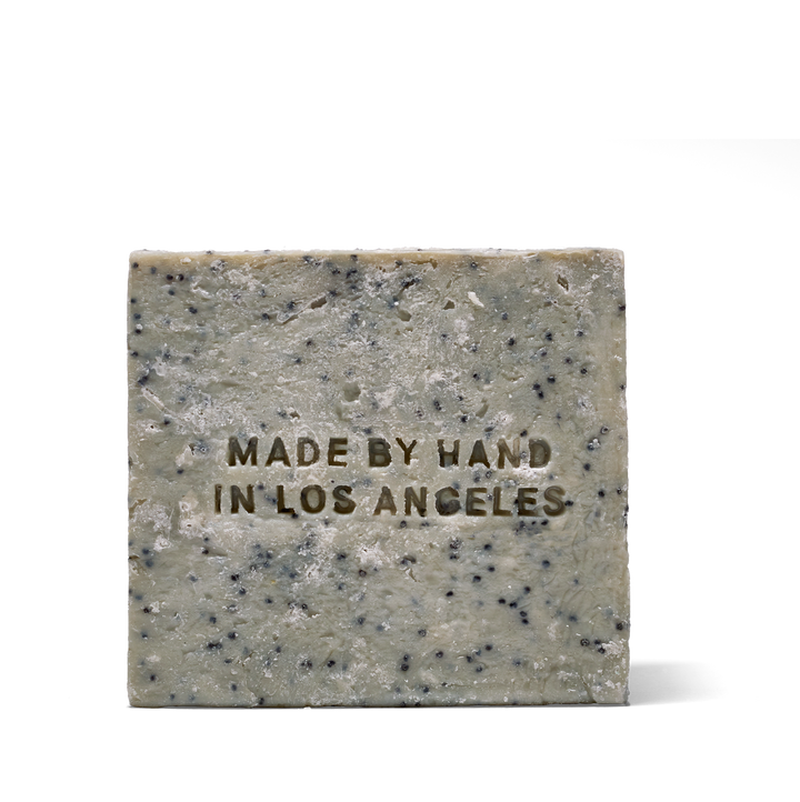 A rectangular bar of speckled soap in a light gray-green tone with dark flecks, featuring the embossed text 'MADE BY HAND IN LOS ANGELES' on the front, set against a plain white background.