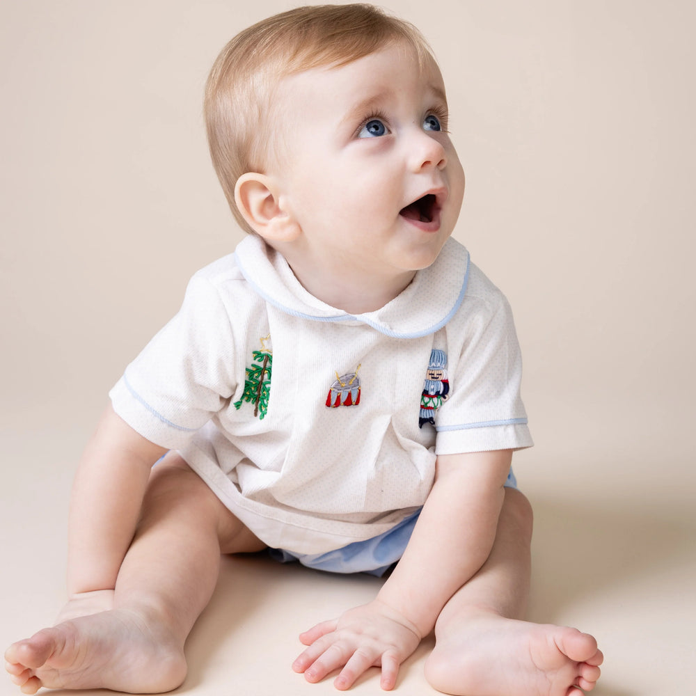 A baby sitting on a beige studio floor wearing a white short-sleeve polo shirt with embroidered holiday motifs on the chest.