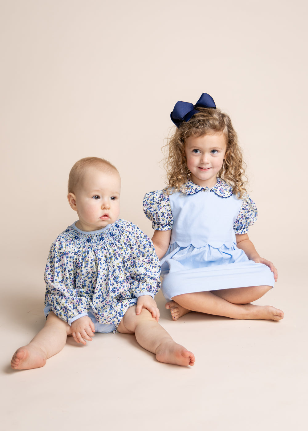 Two young children sitting on a beige studio background. The older child has curly hair and wears a light blue dress with blue floral puff sleeves and a navy bow, while the younger child wears a matching blue floral long-sleeve romper.