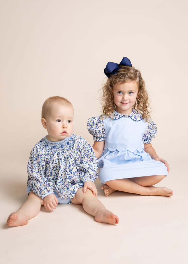 Two young children sitting on a beige studio background. The older child has curly hair and wears a light blue dress with blue floral puff sleeves and a navy bow, while the younger child wears a matching blue floral long-sleeve romper.