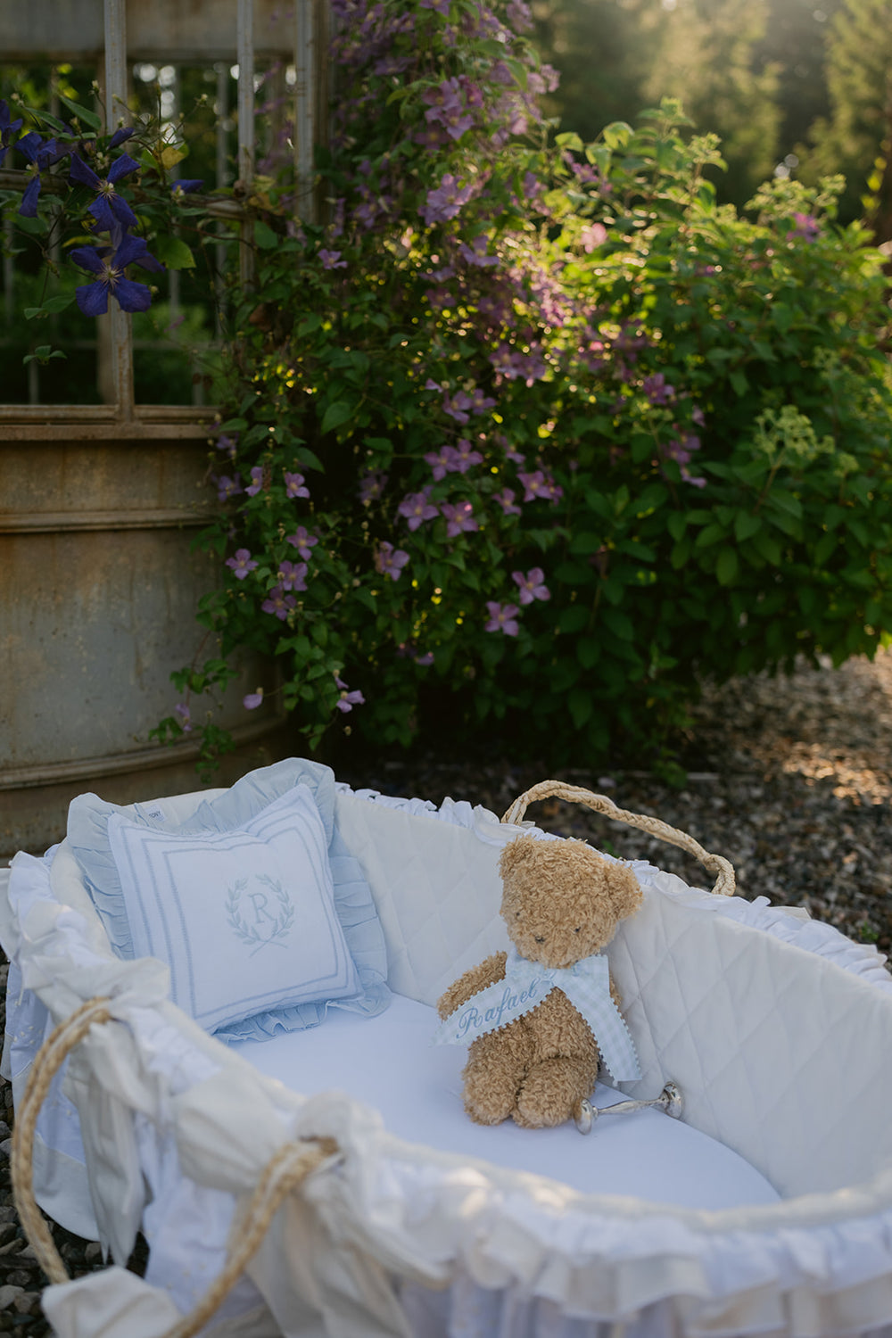 A white ruffled baby bassinet with a teddy bear sitting inside, styled outdoors next to a garden bed with purple flowers and green foliage.