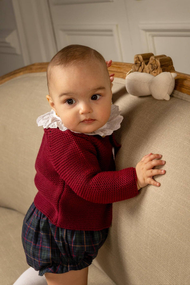 A baby girl wearing a burgundy knit sweater with a white ruffled collar and plaid shorts, standing on a beige fabric sofa.