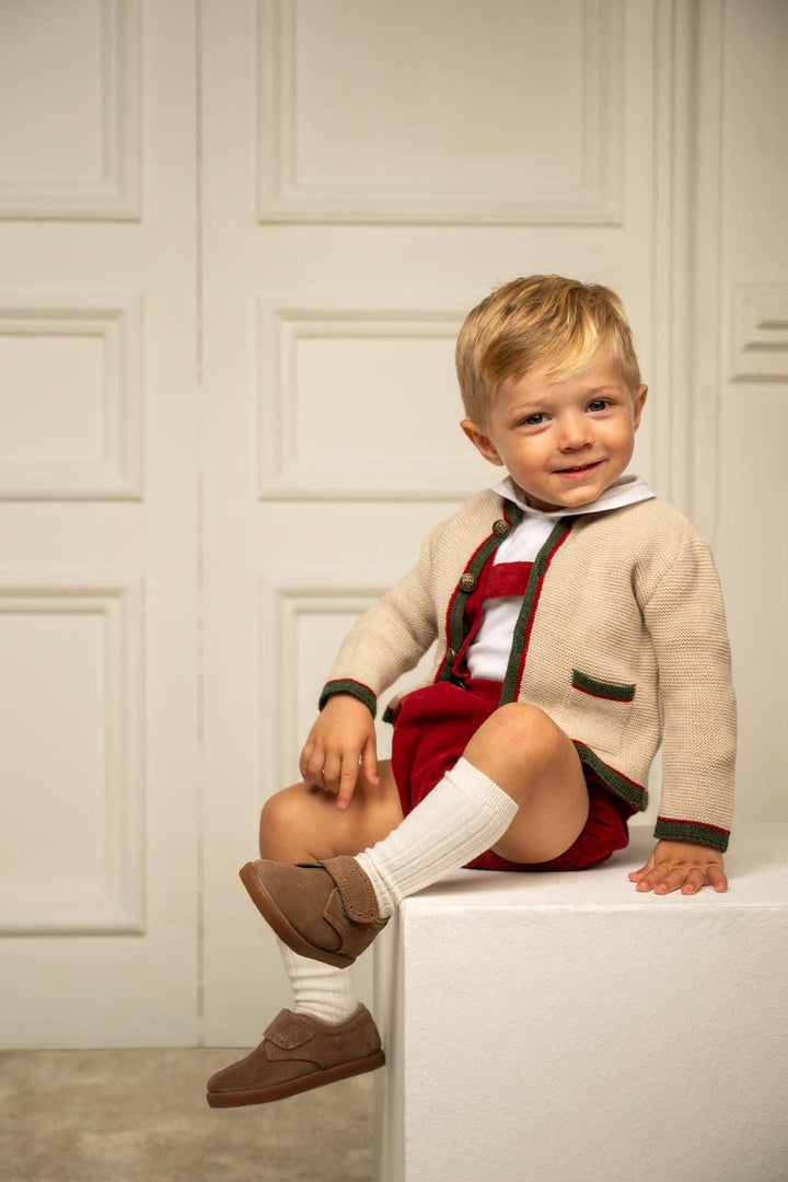 A young toddler boy wearing a cream knit cardigan over a red and white outfit, sitting on a white block in front of a white paneled wall.
