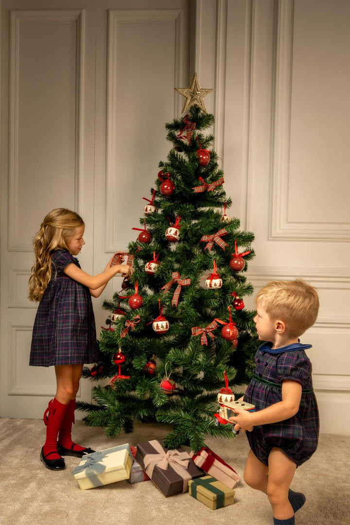 Two children decorating a Christmas tree in a warmly lit room with paneled walls. The tree is adorned with red ornaments, ribbons, and a gold star, with wrapped gifts on the floor below.