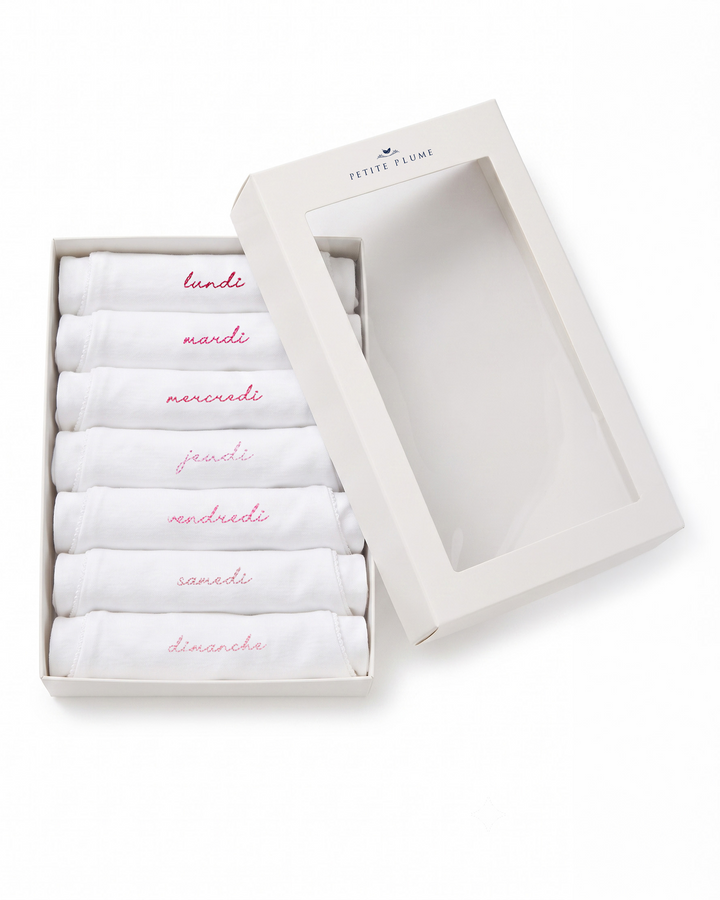 A boxed set of six folded white baby socks or handkerchiefs in a white presentation box with a clear window, shown on a white background. The folded pieces are labeled in pink script with the days of the week in Italian: lunedì, martedì, mercoledì, giovedì, venerdì, sabato, e domenica.