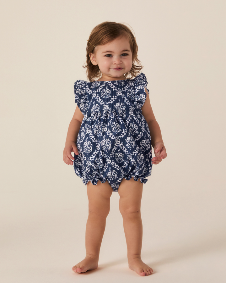 A toddler girl wearing a navy blue and white floral print bubble romper with ruffled shoulders, standing against a solid beige studio background.