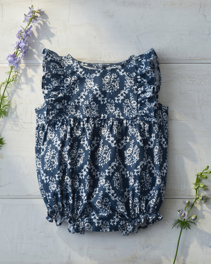 A sleeveless baby romper with a navy blue and white floral damask pattern, featuring ruffled shoulder straps and a smocked waist, laid flat on a white wooden background with decorative purple flowers.