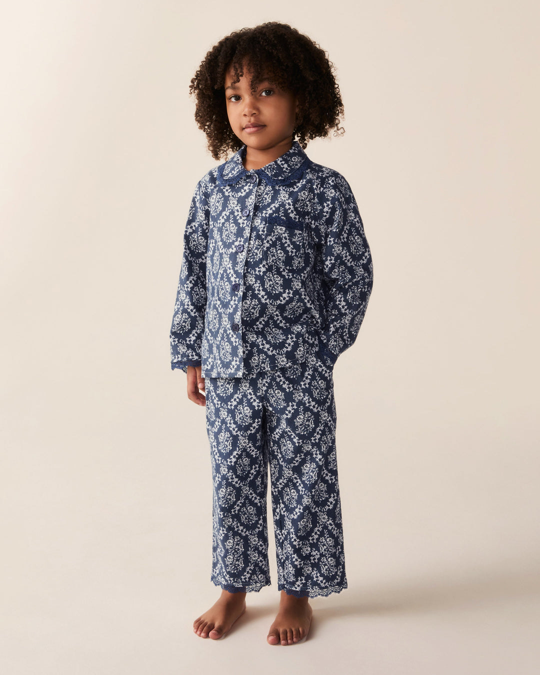 A young child with curly dark hair stands barefoot wearing a matching long-sleeve button-front pajama top and wide-leg pajama pants in a navy blue and white floral pattern, photographed against a plain warm beige studio background.