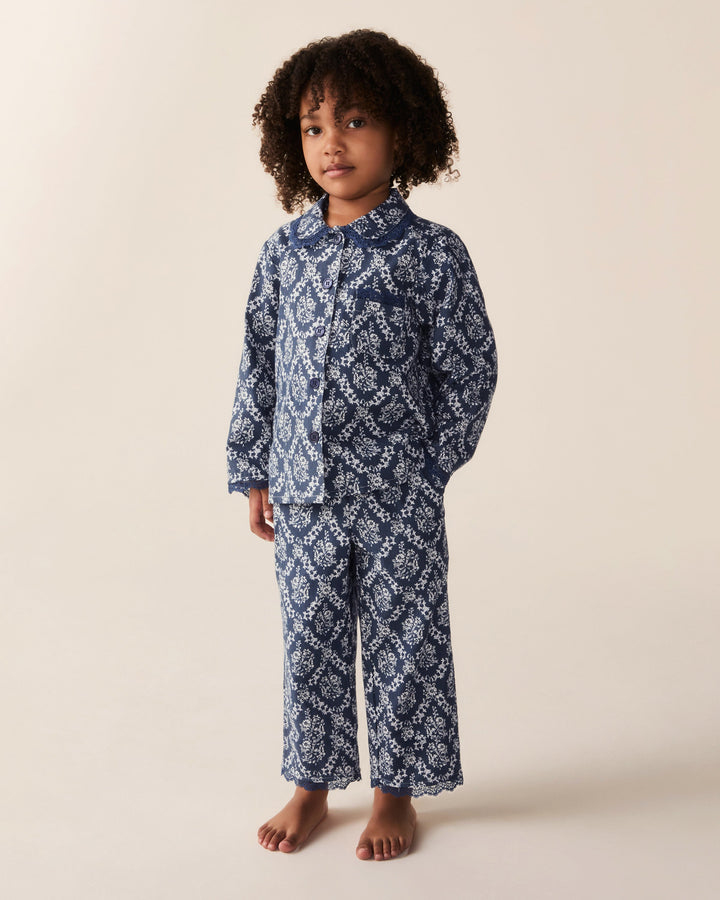 A young child with curly dark hair stands barefoot wearing a matching long-sleeve button-front pajama top and wide-leg pajama pants in a navy blue and white floral pattern, photographed against a plain warm beige studio background.