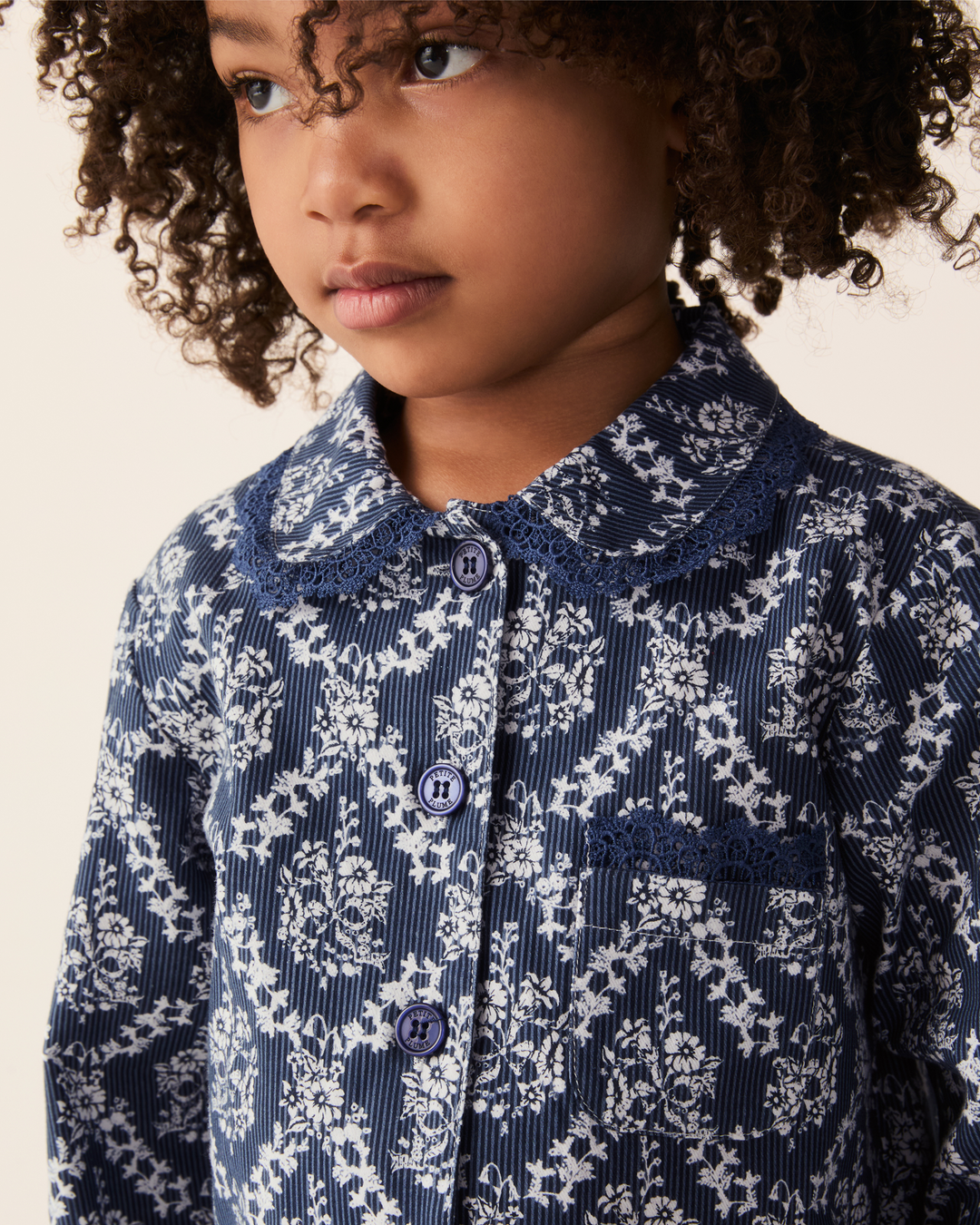 A close-up of a young girl wearing a navy blue and white floral corduroy shirt with a lace Peter Pan collar and matching buttons.