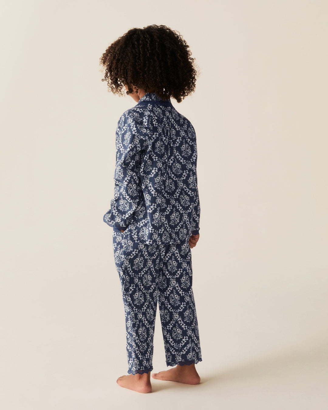 A child standing barefoot from the back, wearing a navy and white patterned long-sleeve pajama set against a plain beige studio background.