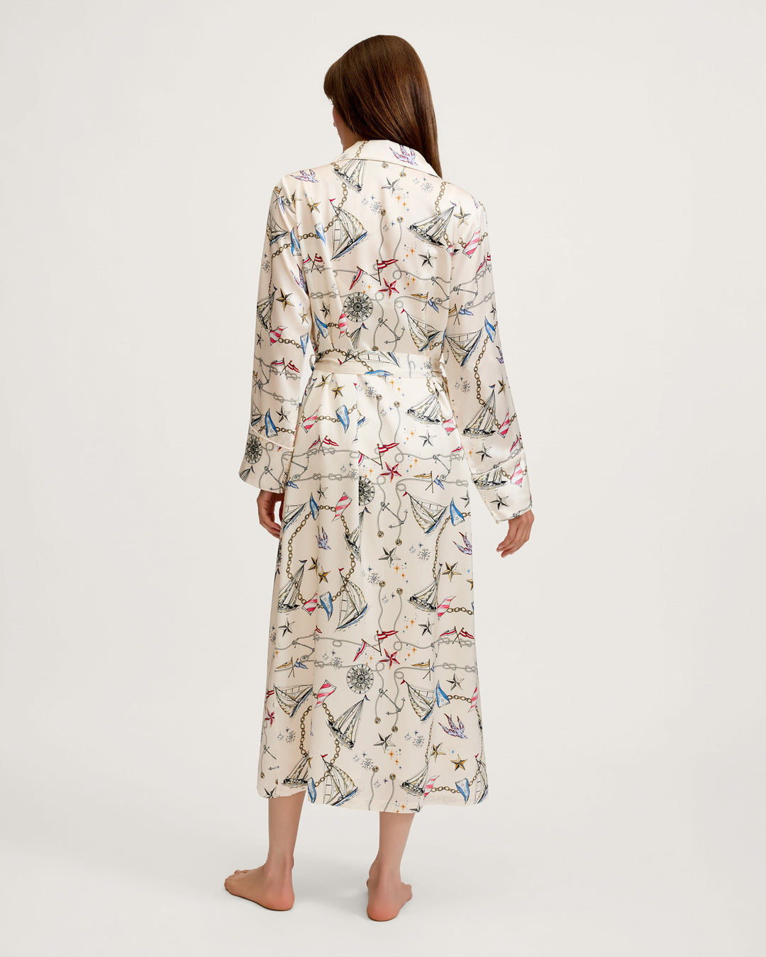 A woman viewed from behind wearing a long-sleeve belted robe with an all-over abstract print in cream, blue, red, and black, standing barefoot against a plain light gray studio background.