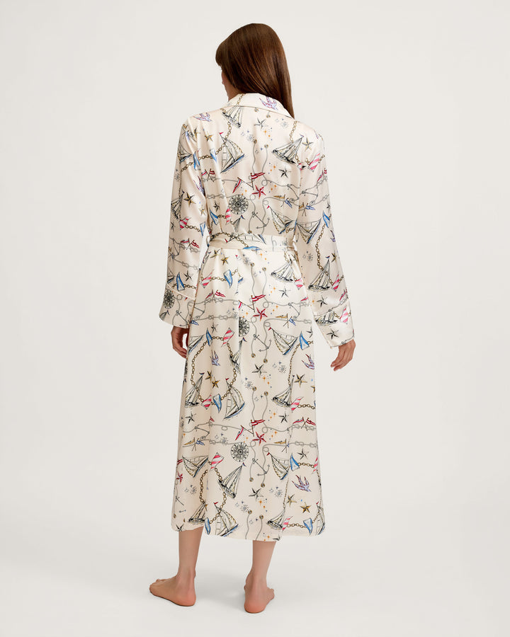 A woman viewed from behind wearing a long-sleeve belted robe with an all-over abstract print in cream, blue, red, and black, standing barefoot against a plain light gray studio background.