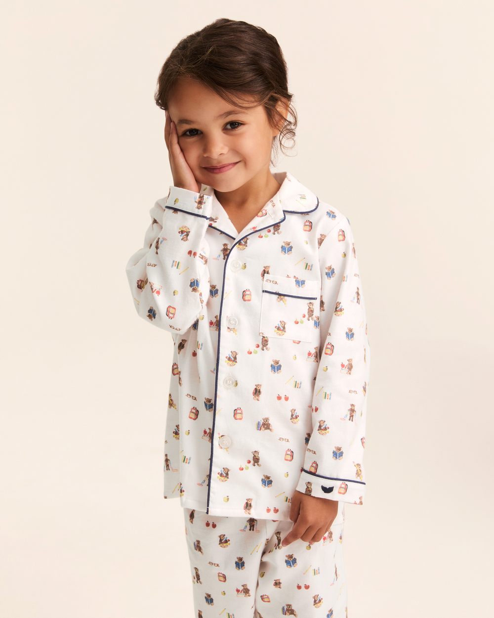 A young girl wearing a white long-sleeve pajama set with a colorful cartoon character print and navy piping, posing against a solid light beige background.