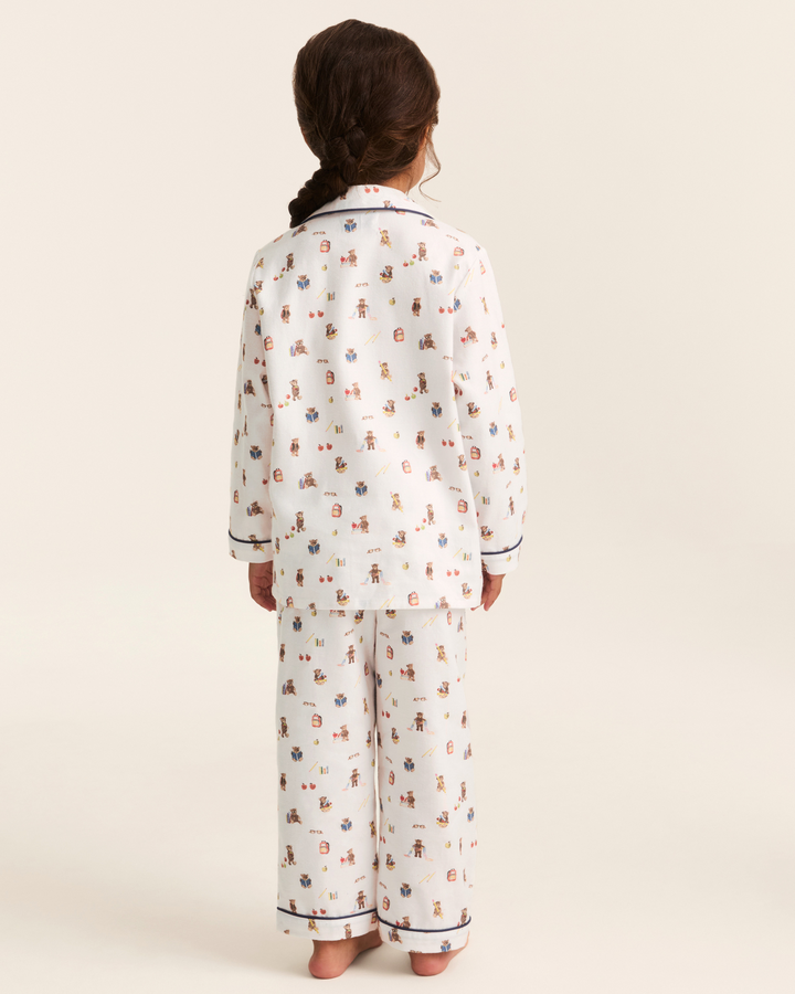 A child shown from the back wearing a matching pajama set with a white base and a small multicolor pattern of cartoon figures, photographed against a plain warm off-white studio background.