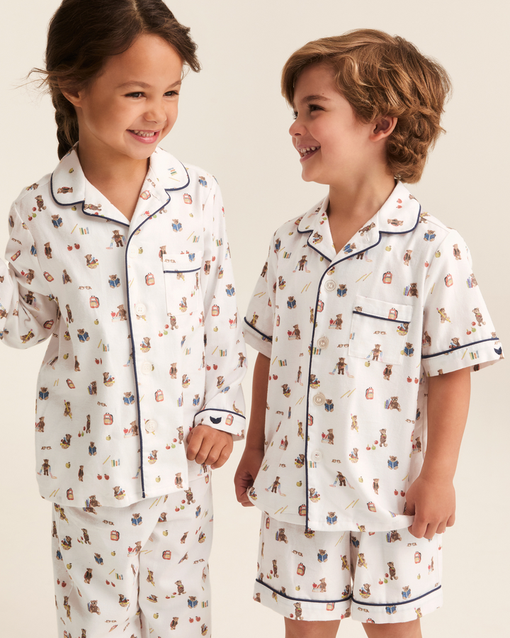Two children wearing matching short-sleeve pajama sets with a white base and a colorful cartoon print, standing against a plain light beige background.