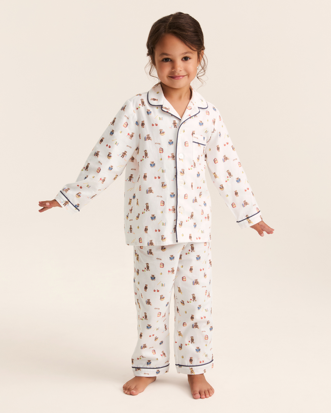 A young girl stands barefoot against a warm off-white studio background wearing a two-piece pajama set. The pajamas are white with a small scattered print in muted red, blue, and brown, and have dark navy piping along the collar, cuffs, and hem.