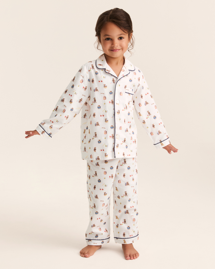 A young girl stands barefoot against a warm off-white studio background wearing a two-piece pajama set. The pajamas are white with a small scattered print in muted red, blue, and brown, and have dark navy piping along the collar, cuffs, and hem.