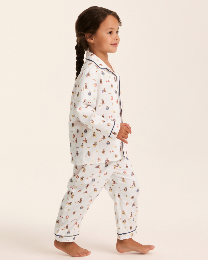 A young girl with her hair in a braid is shown in profile wearing a two-piece pajama set. The pajamas are white with a small, colorful teddy bear pattern and dark blue piping on the collar, placket, cuffs, and hem.