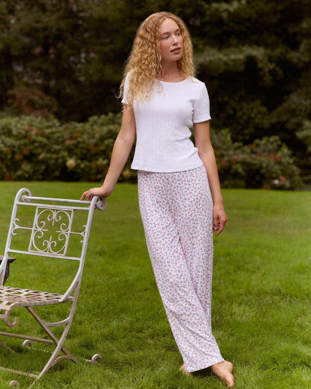 Woman in white top and patterned pants standing outdoors with a chair.