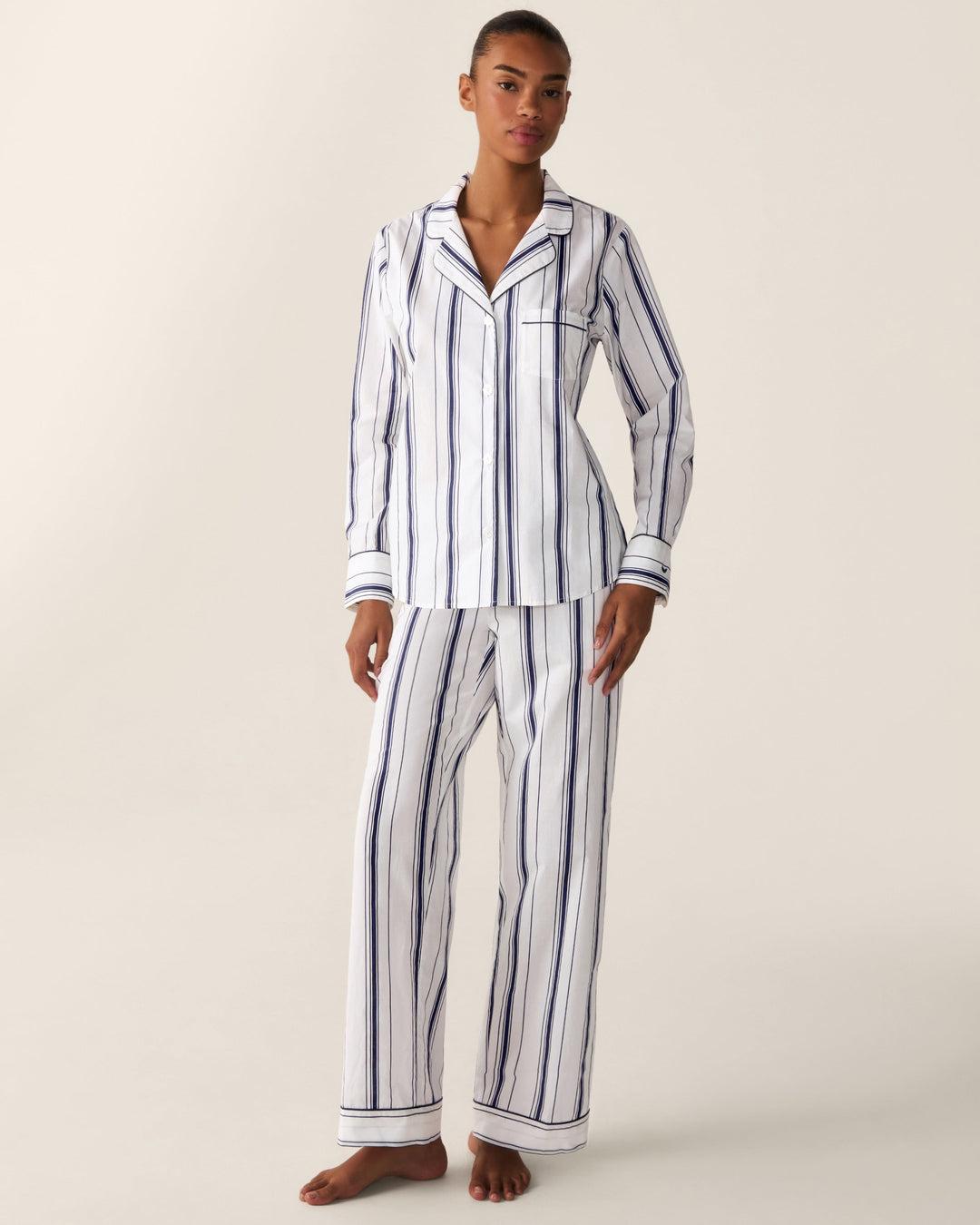 A woman modeling a matching two-piece pajama set with vertical navy blue and white stripes, consisting of a long-sleeve button-up top and wide-leg pants, against a plain beige studio background.