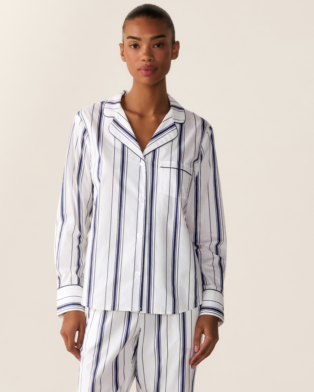 A model wearing a white long-sleeve pajama shirt and matching pants with vertical navy blue stripes, photographed against a solid light beige background.