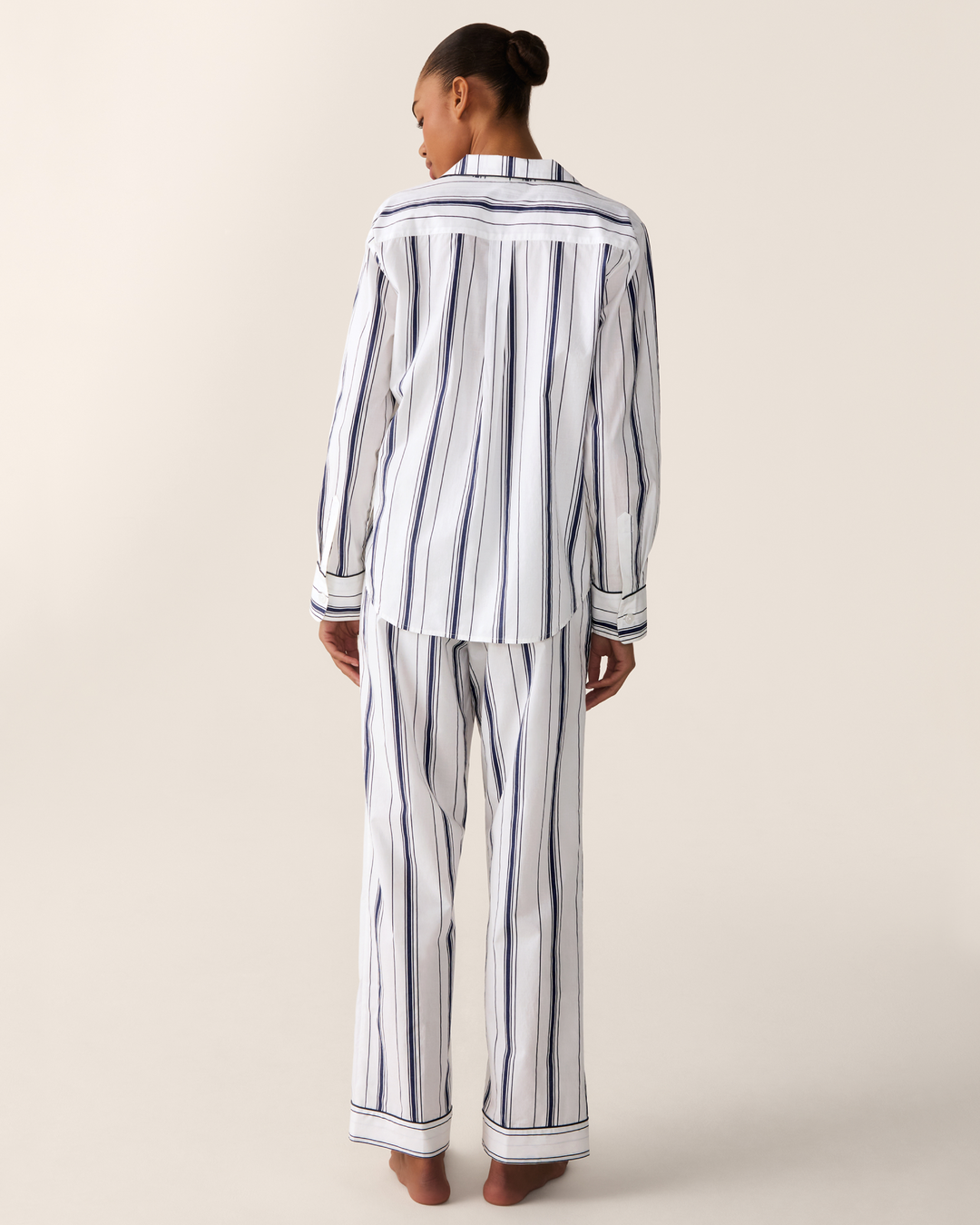 A woman shown from the back wearing a matching long-sleeve button-up pajama shirt and wide-leg pajama pants set, both featuring vertical navy blue stripes on a white background, against a solid light beige studio backdrop.