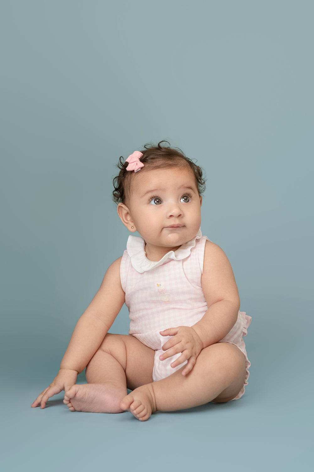 Baby sitting on a plain background wearing a light pink outfit with a bow.