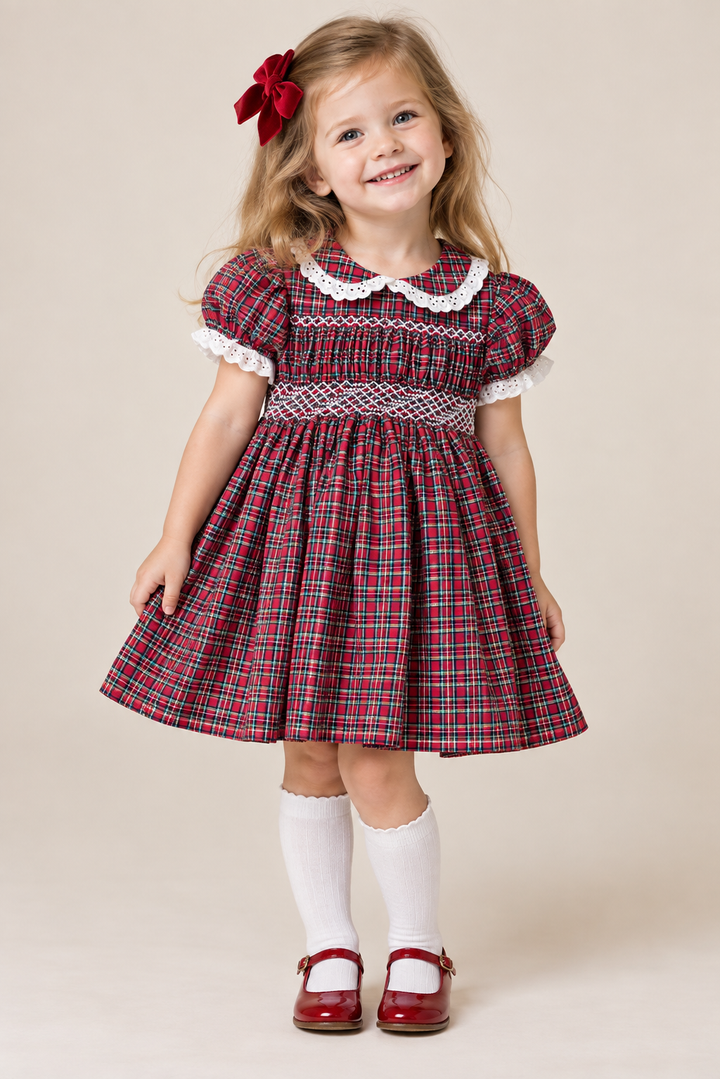A young girl with blonde hair wearing a red, green, and blue plaid dress with white lace trim, white knee-high socks, and red Mary Jane shoes, standing against a plain beige background.